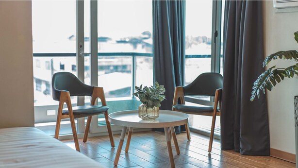 A hotel room with a bed, two chairs, a small table with a plant, and large windows with curtains. - Henia Hotel (Dumaguete)