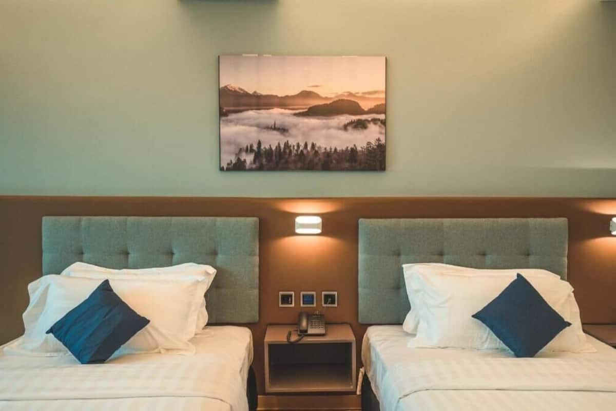 Quarto de hotel com duas camas, uma mesa de cabeceira com telefone e um quadro pendurado na parede retratando uma paisagem montanhosa.
