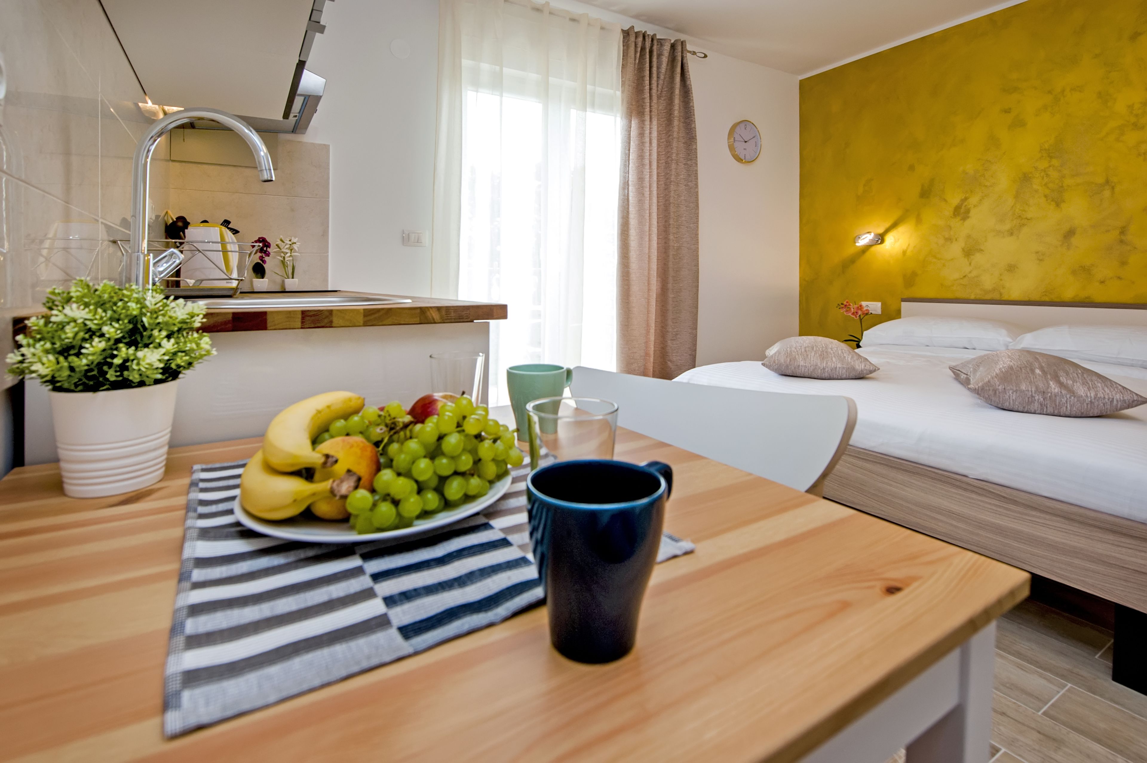 A hotel room with a bed, a bedside table with a plate of fruit and a mug, a small potted plant, and a window with curtains.