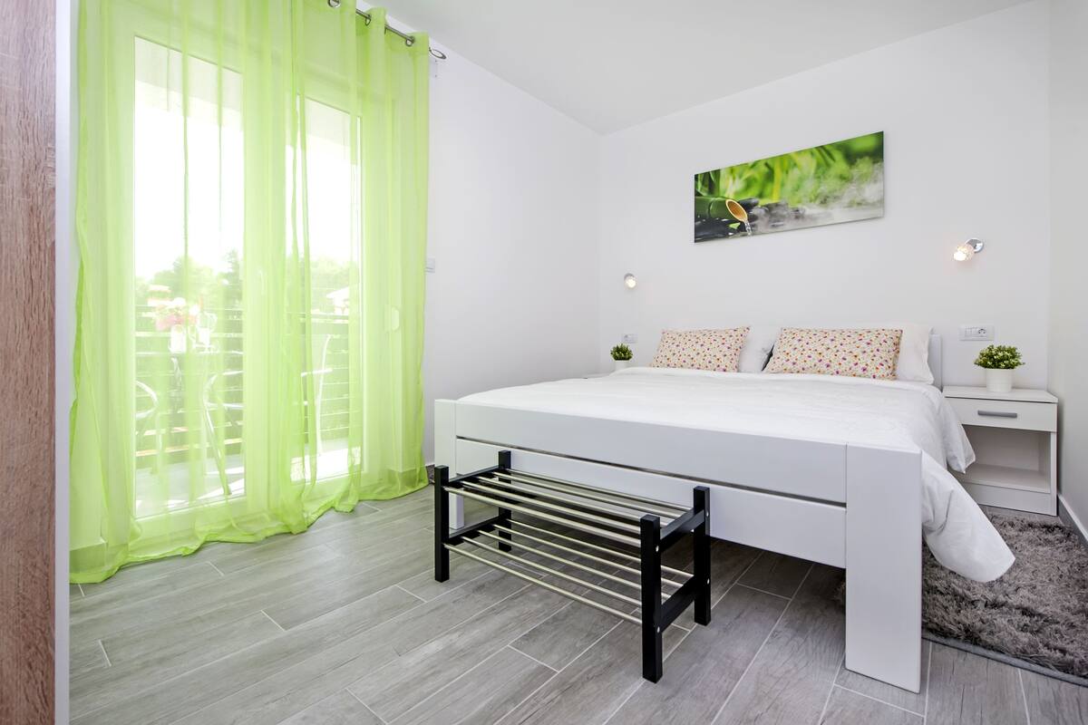 Quarto moderno com cama, varal de roupas, mesa de cabeceira e janela com cortinas leves.