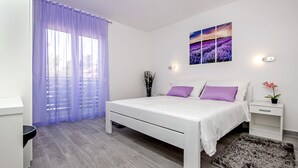 A modern bedroom with a white bed, purple pillows, a nightstand with a plant, and a large window with purple curtains. - Apartment and Studio Tereza (Rovinj)