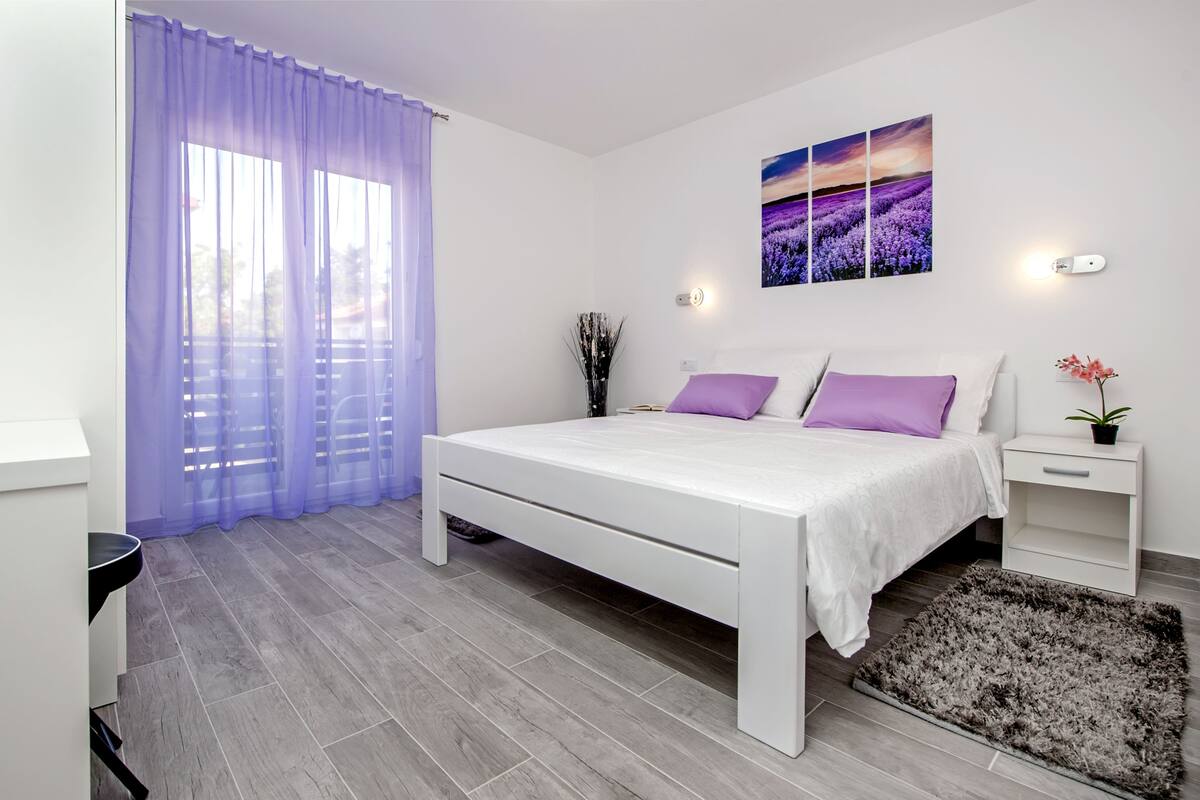 Um quarto moderno com uma cama branca, almofadas roxas, uma mesinha de cabeceira com uma planta e uma janela grande com cortinas roxas.