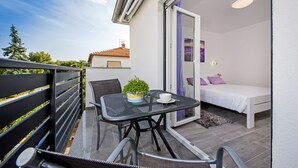 A balcony with a table and chairs, a bed with a white mattress and purple pillows, and a view of the sky and trees. - Apartment and Studio Tereza (Rovinj)