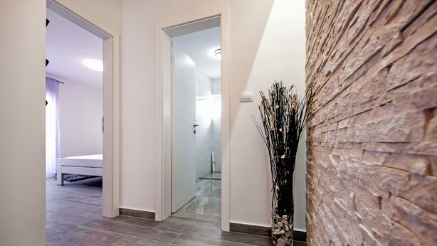 A hallway with a bed in the background, a stone wall, and a vase with dried plants. - Apartment and Studio Tereza (Rovinj)