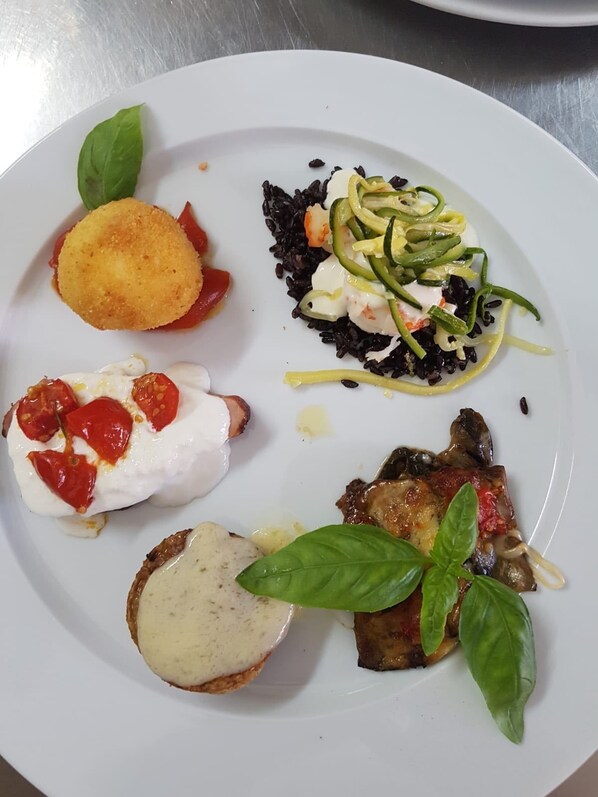Un plato con diferentes alimentos, incluyendo un bocado empanizado, una ensalada de arroz negro, un trozo de carne con albahaca y una rebanada de pan con cobertura cremosa.