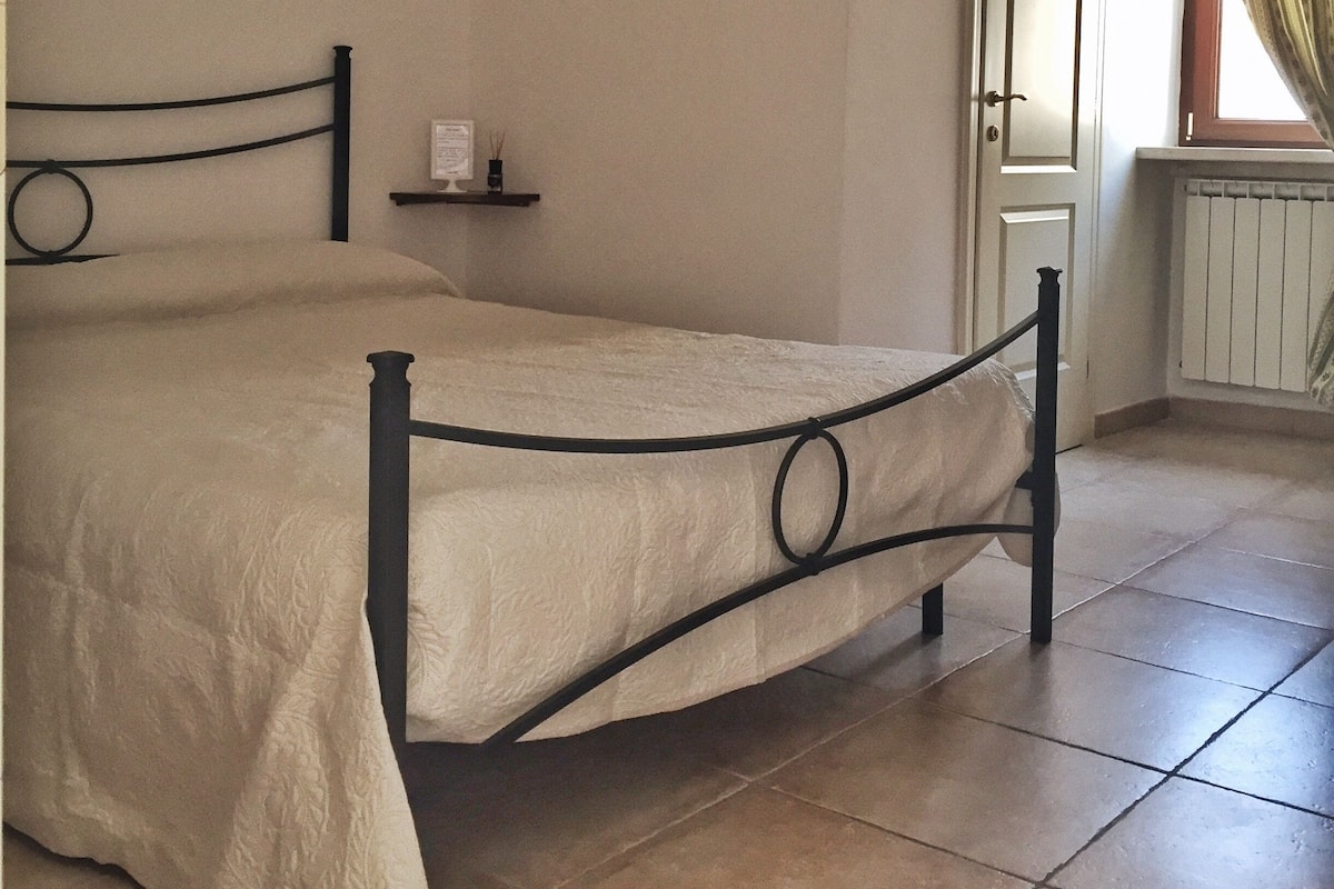 A bedroom with a double bed, a bedside table, a wall clock, and a window with curtains.