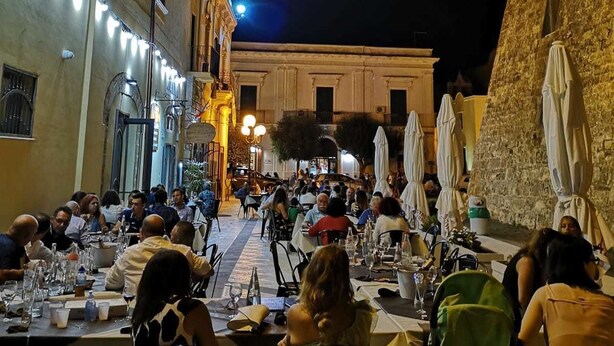 Un espacio de comedor al aire libre con mesas y sillas, rodeado de edificios con ventanas iluminadas y terrazas.