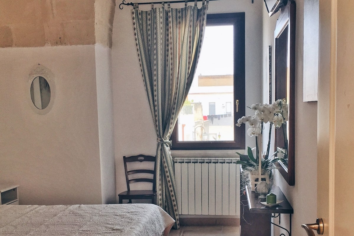 A bedroom with a bed, a chair, a radiator, a window with curtains, and a TV mounted on the wall.