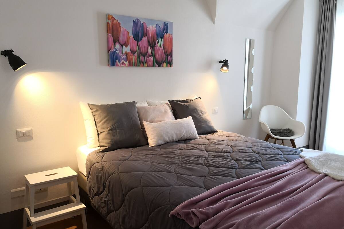 Quarto com cama, almofadas, mesa de cabeceira, cadeira, pintura e arandelas.