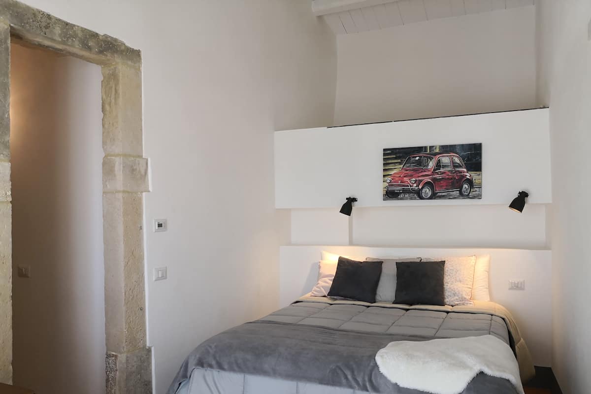 Quarto com uma cama, cabeceira, um quadro na parede e uma pequena luminária preta.
