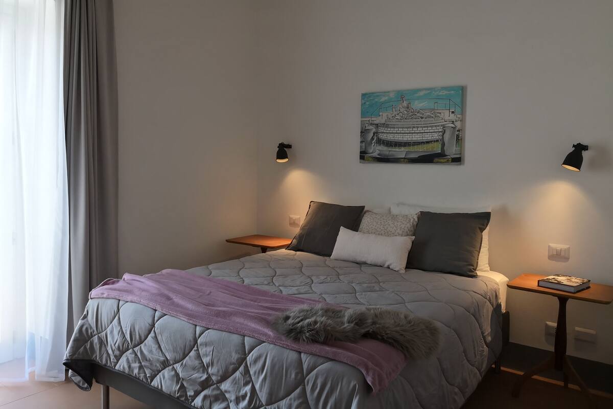 Quarto com cama, mesa de cabeceira, luminária na parede e um quadro.
