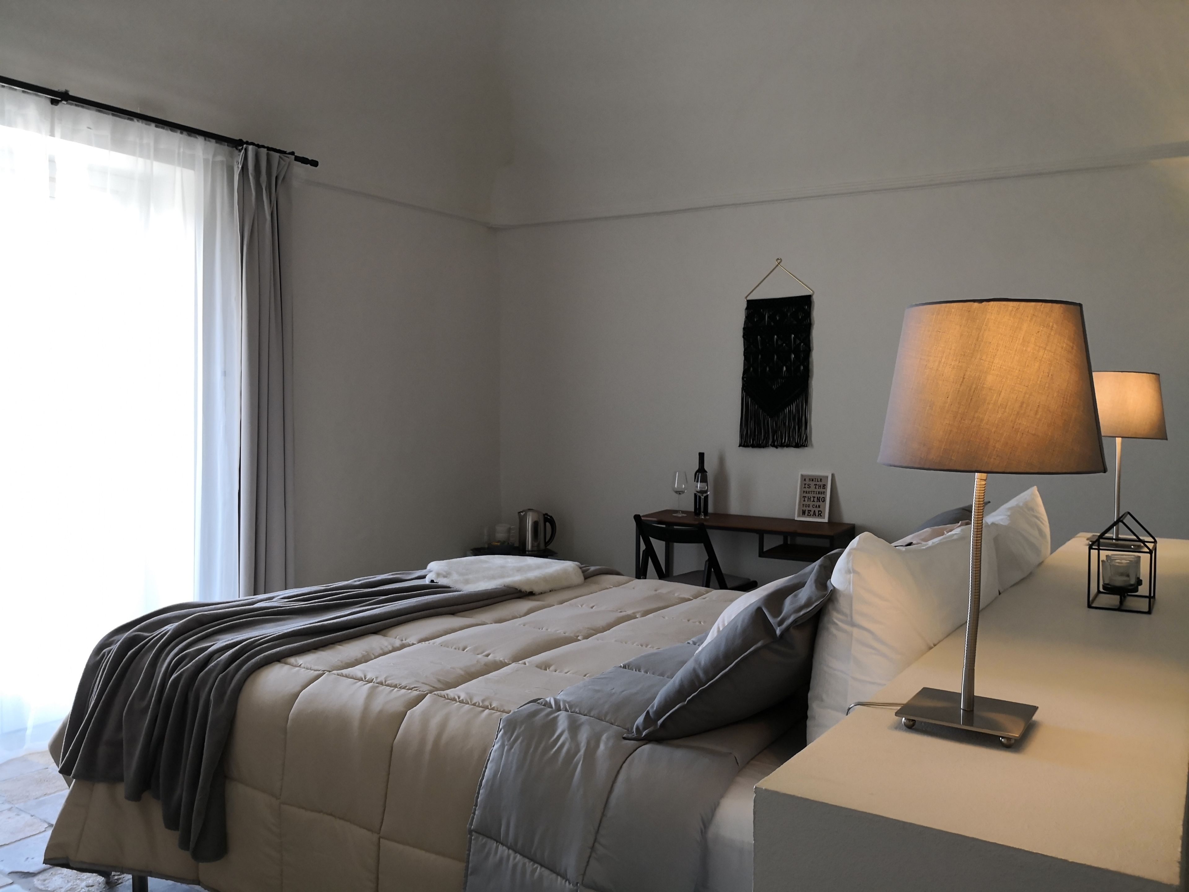 A neatly arranged hotel room with a bed, bedside table, lamp, and a small desk with a kettle and wine glasses.