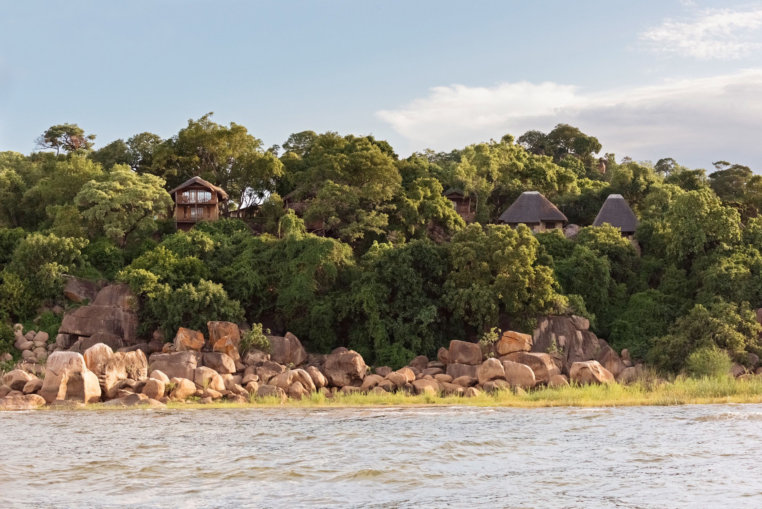 A secluded lodge with multiple thatched-roof cabins nestled among dense trees and large rocks by a calm river.