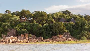 A secluded lodge with multiple thatched-roof cabins nestled among dense trees and large rocks by a calm river.