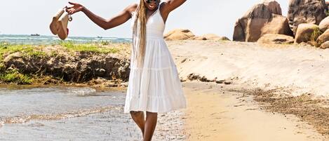 A woman in a white dress standing on a sandy beach with her arms raised, holding a pair of sandals.