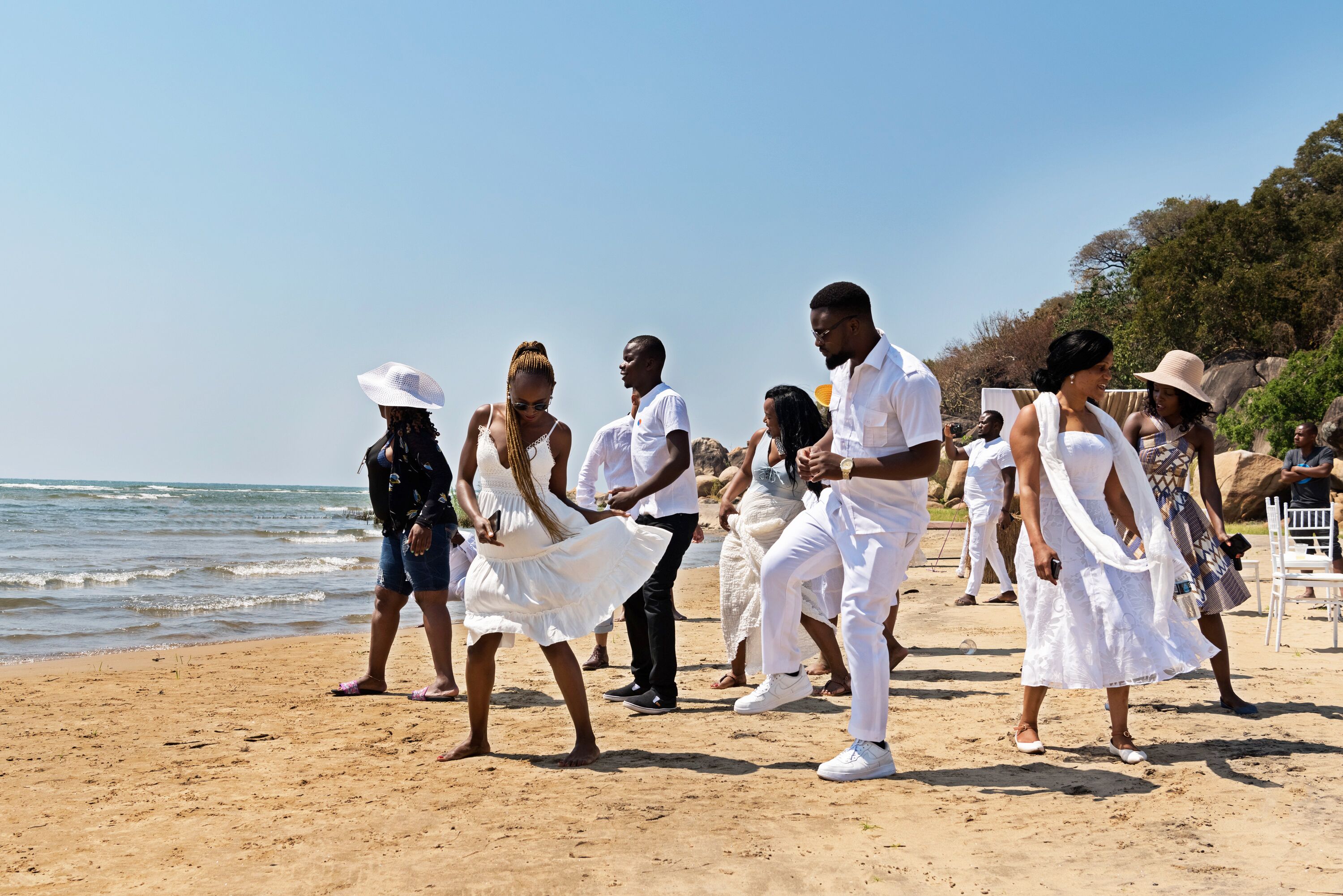 Un groupe de personnes se trouve sur une plage de sable, certaines d’entre elles portent des vêtements blancs, et l’océan est visible en arrière-plan.