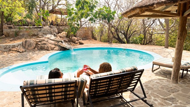 Two lounge chairs by a swimming pool with a stone patio and a thatched roof structure in the background. - Safari Beach Lodge (Senga)