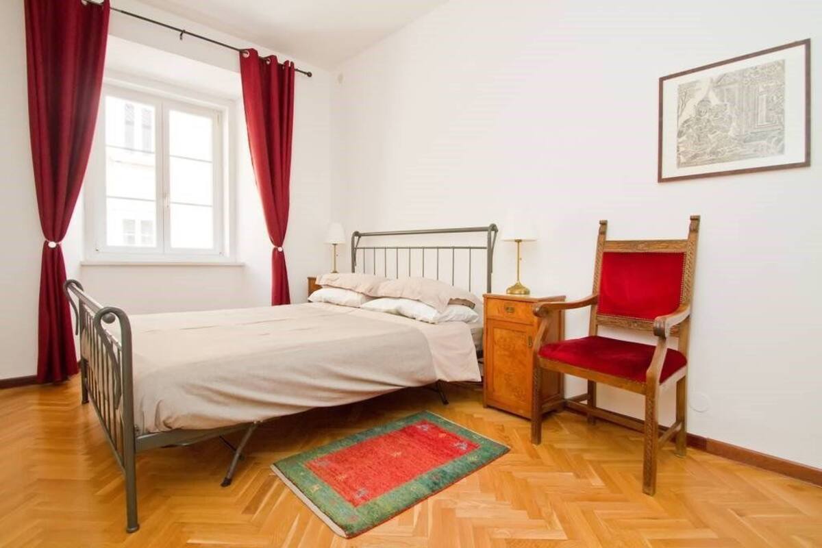 Un dormitorio con estructura de cama metálica, mesita de noche de madera, una silla roja, un tapete rojo y verde, y un cuadro en la pared.