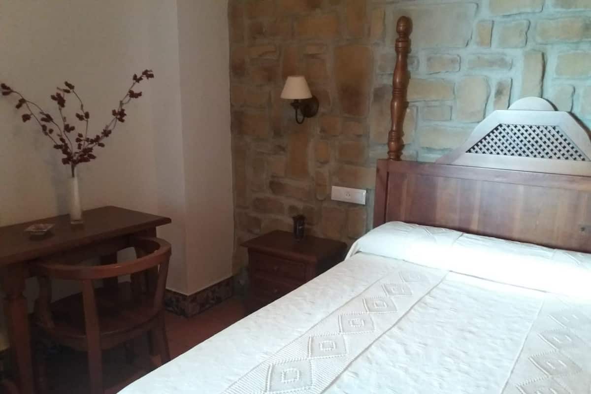 A bedroom with a wooden bed, a nightstand, a desk, and a lamp against a stone wall.