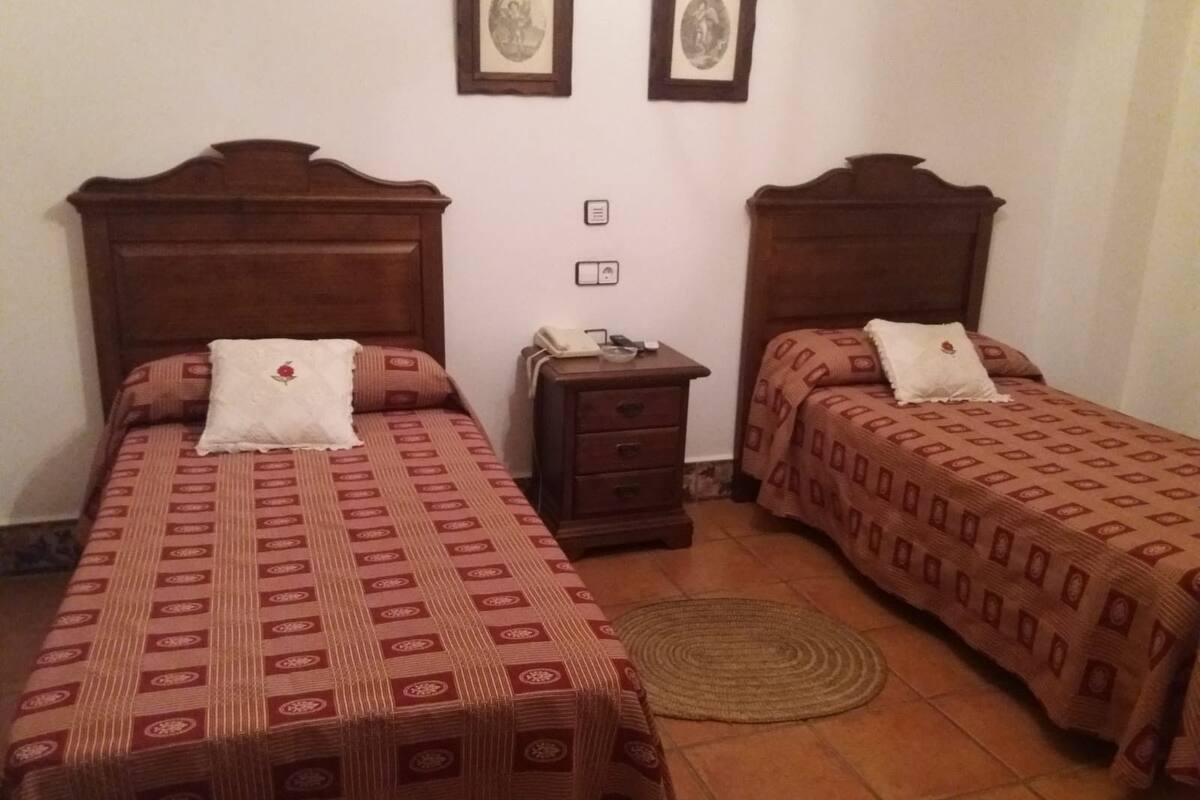 A room with two beds, each with a patterned bedspread and a white pillow. There is a small wooden nightstand with a telephone between the beds.