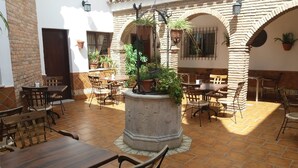 Un patio al aire libre con varias mesas y sillas de madera, rodeado de muros y arcos de ladrillo, con plantas en macetas y faroles colgantes.