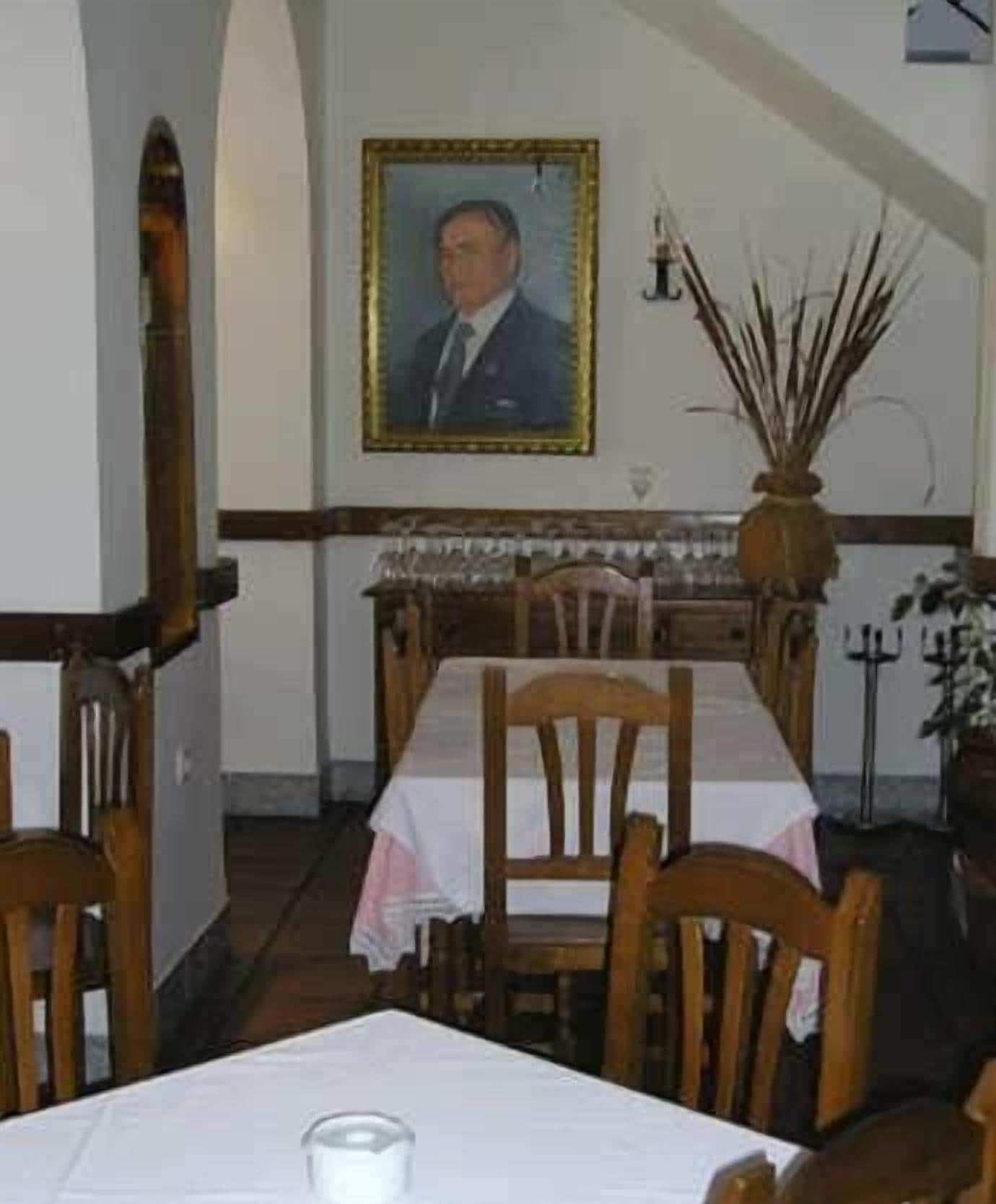 Un comedor con una mesa para dos, sillas de madera, un retrato enmarcado en la pared y un jarrón con plantas secas.
