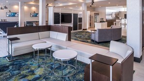A modern hotel lobby with a sectional sofa, two round tables, a kitchen area, and a large TV screen.