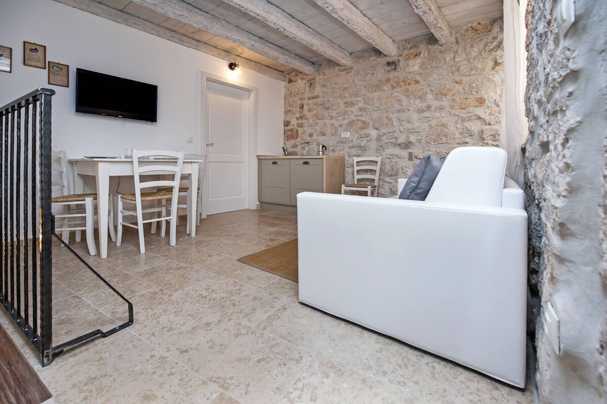 A modern living room with a white sofa, a dining table with chairs, a kitchen area, and a stone wall.