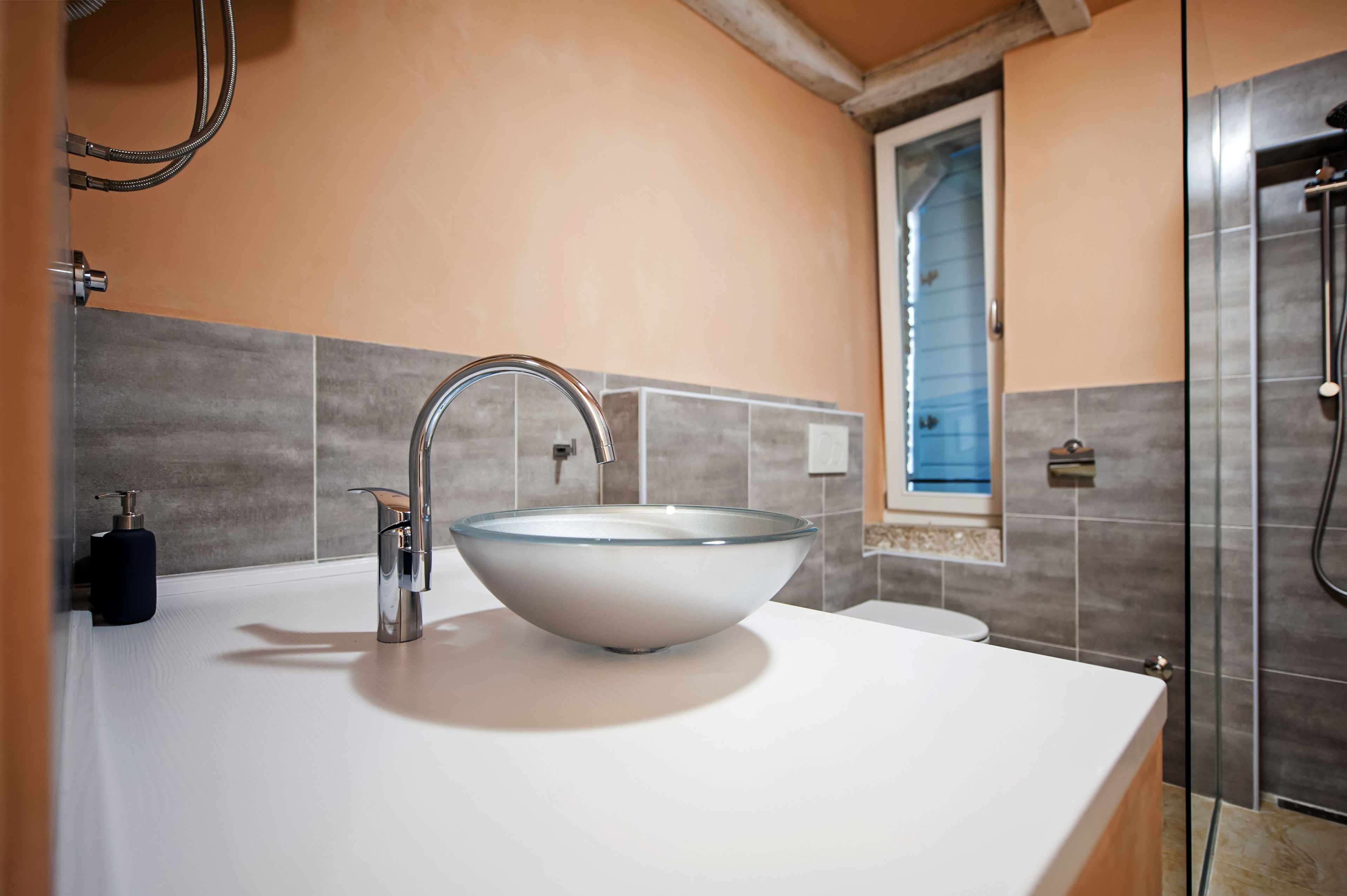 Une salle de bain avec un lavabo en forme de bol en verre, une douchette à main et une fenêtre donnant sur l’extérieur.