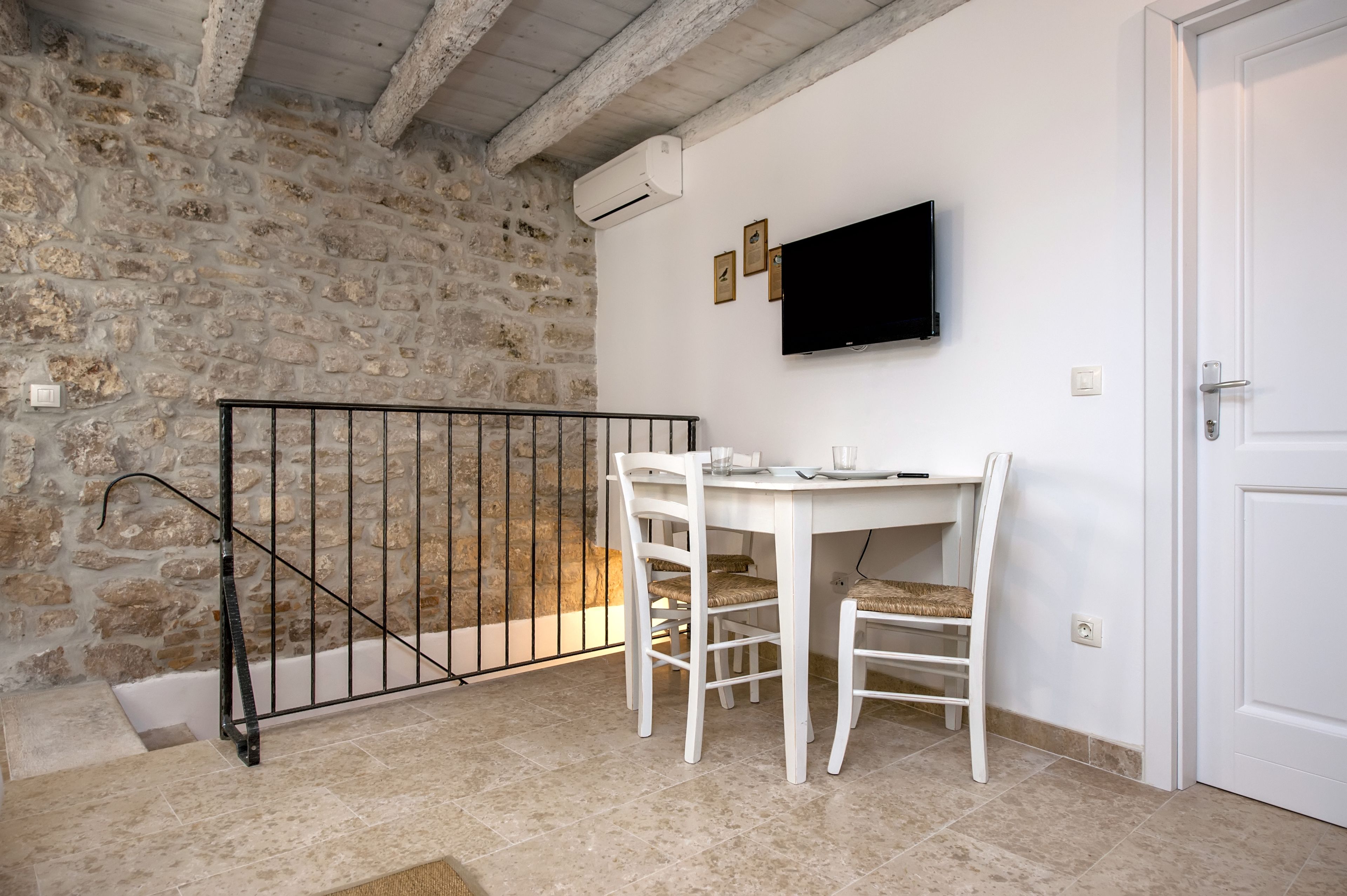 Une pièce avec un mur en pierre, une porte blanche, un téléviseur fixé au mur et une table avec des chaises.