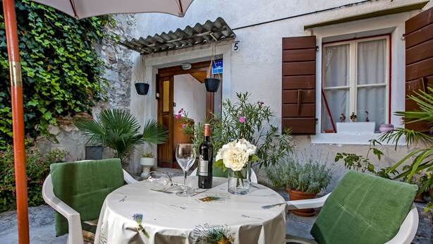 Un área de comedor al aire libre con una mesa dispuesta para dos personas, rodeada de macetas con plantas y un edificio rústico al fondo.