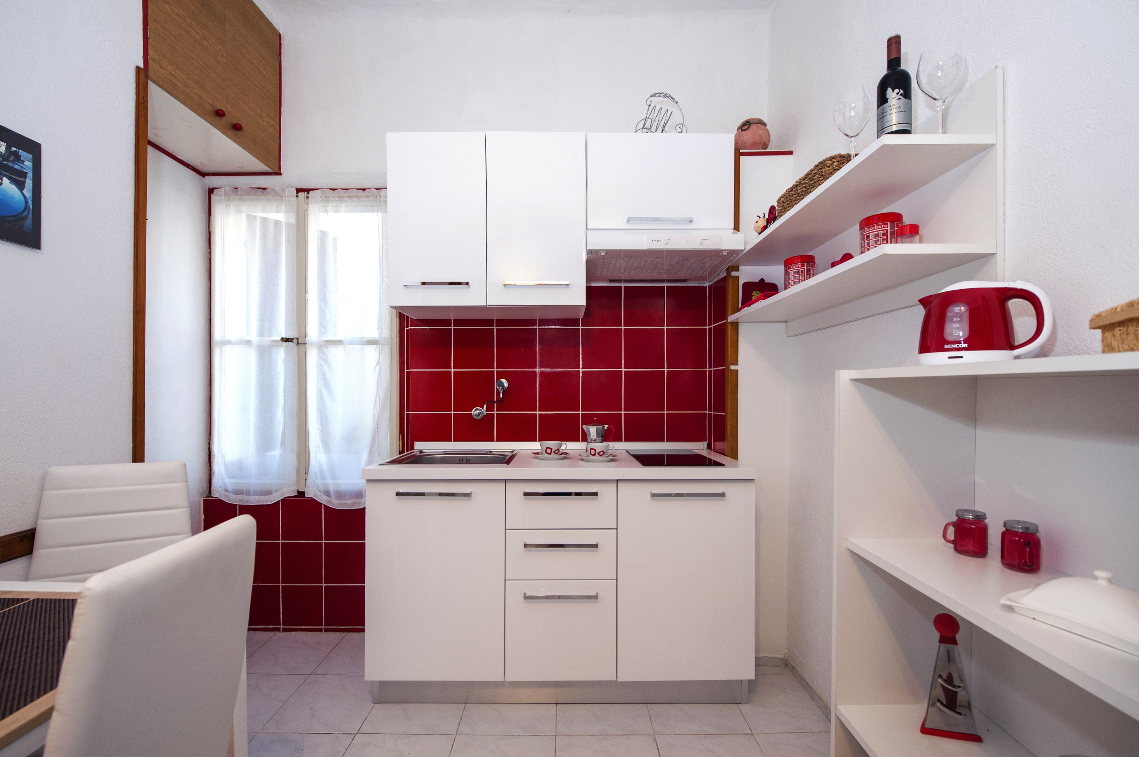 A compact kitchen with white cabinets, a red tiled backsplash, and a small dining area with a chair.