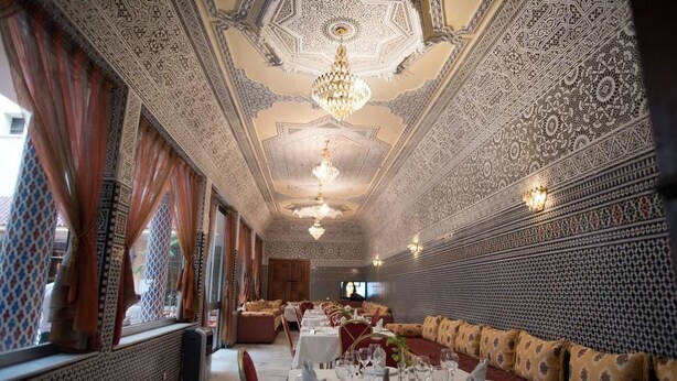 Un comedor con elaborados diseños en el techo, candelabros y paredes con motivos geométricos. Las mesas están dispuestas con manteles blancos y sillas rojas.