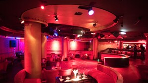 Un área de estar tenue con iluminación ambiental roja, un conjunto de asientos en el centro y un bar al fondo.