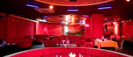 Un restaurante con iluminación roja, que incluye mesas, cabinas, una barra y un escenario al fondo.