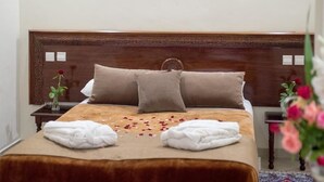 A neatly made bed with a brown headboard, two pillows, and a beige bedspread. There are two white towels folded on the bed and a vase with a single red rose on a nightstand beside the bed.