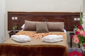 A neatly made bed with a brown headboard, two pillows, and a beige bedspread. There are two white towels folded on the bed and a vase with a single red rose on a nightstand beside the bed.