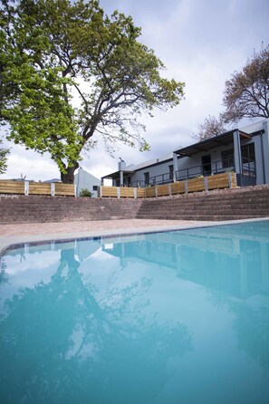 A swimming pool with a clear blue water surface, surrounded by a brick wall and a wooden deck. In the background, there is a modern building with large windows and a flat roof. - Mont Angelis Retreat (Stellenbosch)
