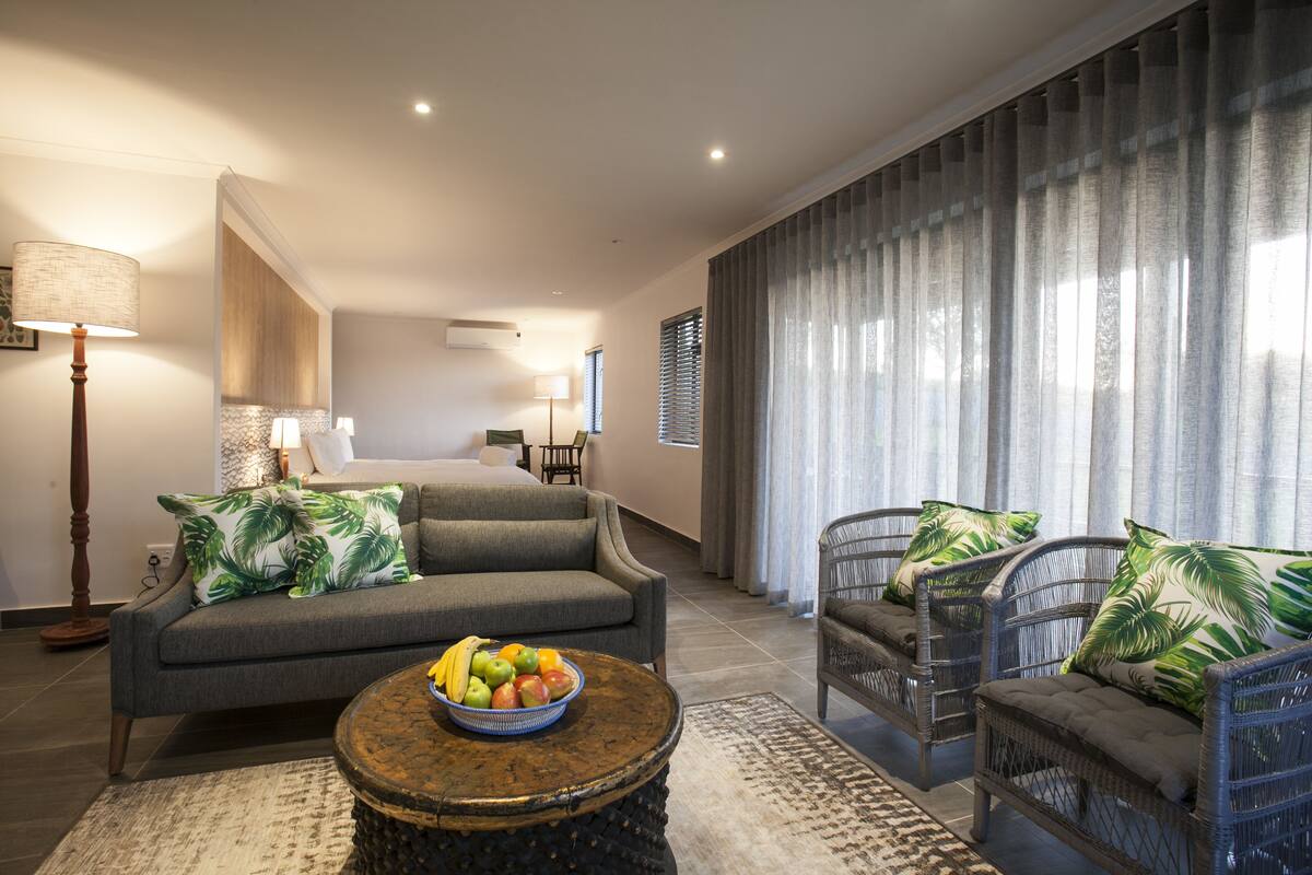A modern living room with a grey sofa, wicker chairs, a round coffee table with fruit, and a large window with curtains.