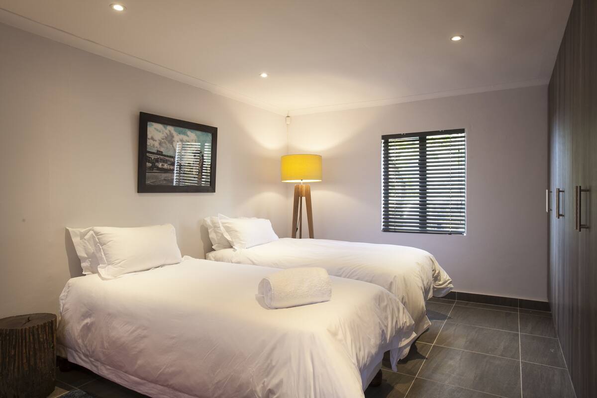 A hotel room with two beds, a lamp, a framed picture, and a window with blinds.