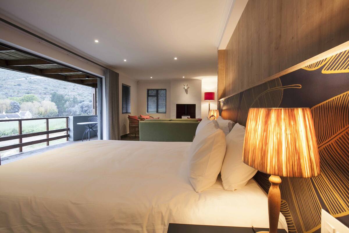 A large bed with white linens, a wooden headboard, and a bedside lamp. There is a view of a balcony with a railing and a table.
