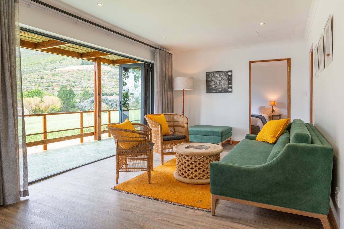 A spacious living area with a green sofa, wicker chairs, and a round side table. A large window offers a view of a hillside.