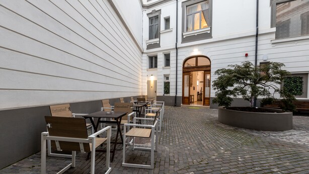 Un espace extérieur aménagé avec des tables et des chaises, un arbre en pot et l’entrée d’un bâtiment.
