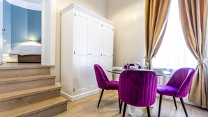 Une chambre d’hôtel moderne avec un escalier en bois, une armoire blanche, une table à manger ronde entourée de chaises violettes et une fenêtre avec des rideaux.