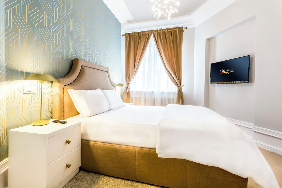 Habitación de hotel con cama, mesita de noche, televisor y ventana con cortinas.