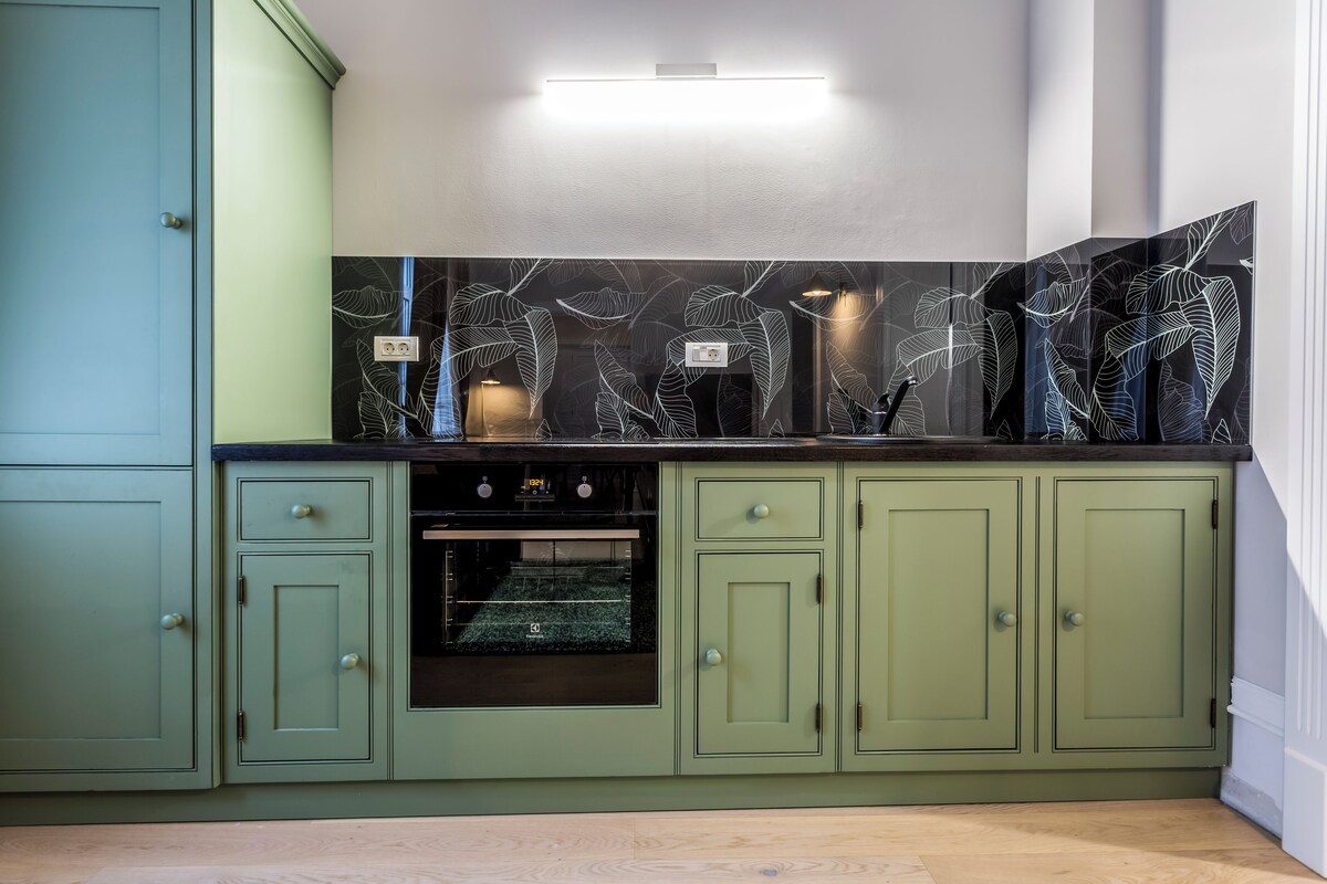 Una cocina con armarios verdes, un salpicadero negro con motivos de hojas y un horno empotrado.