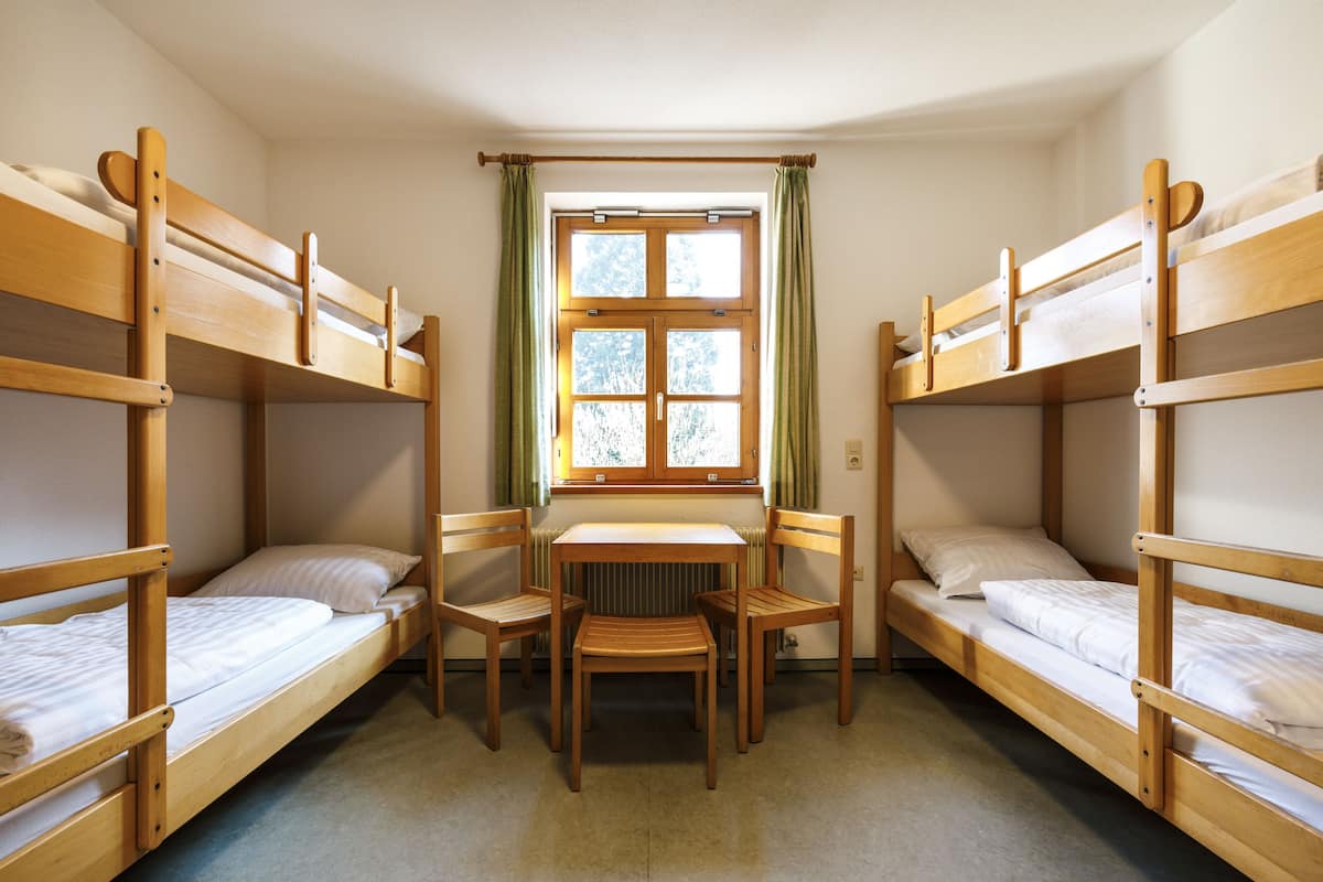 Een slaapzaal met stapelbedden, een tafel, stoelen en een raam met gordijnen.