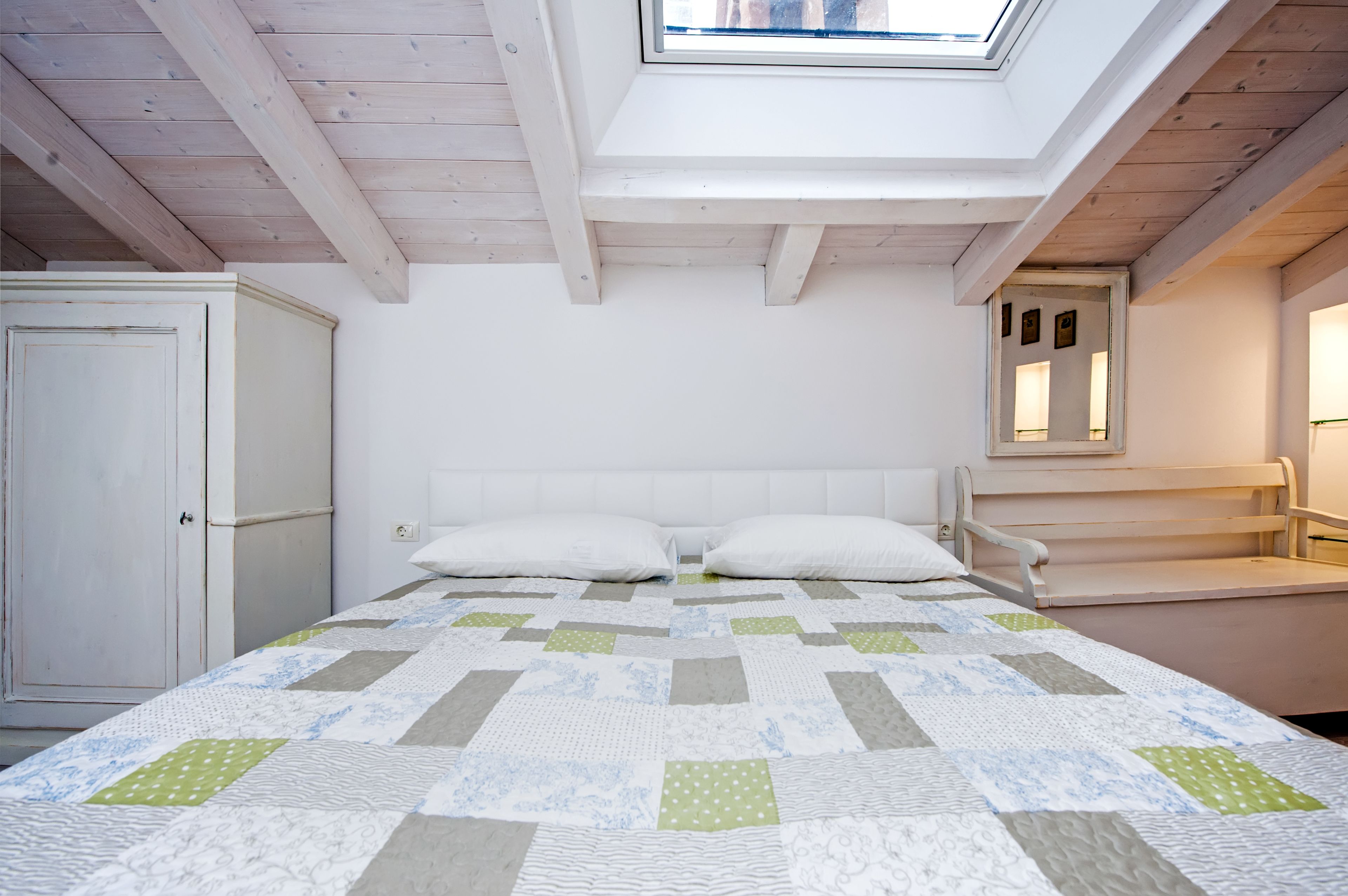 A bedroom with a skylight, a bed with a quilt, a wardrobe, and a bench.