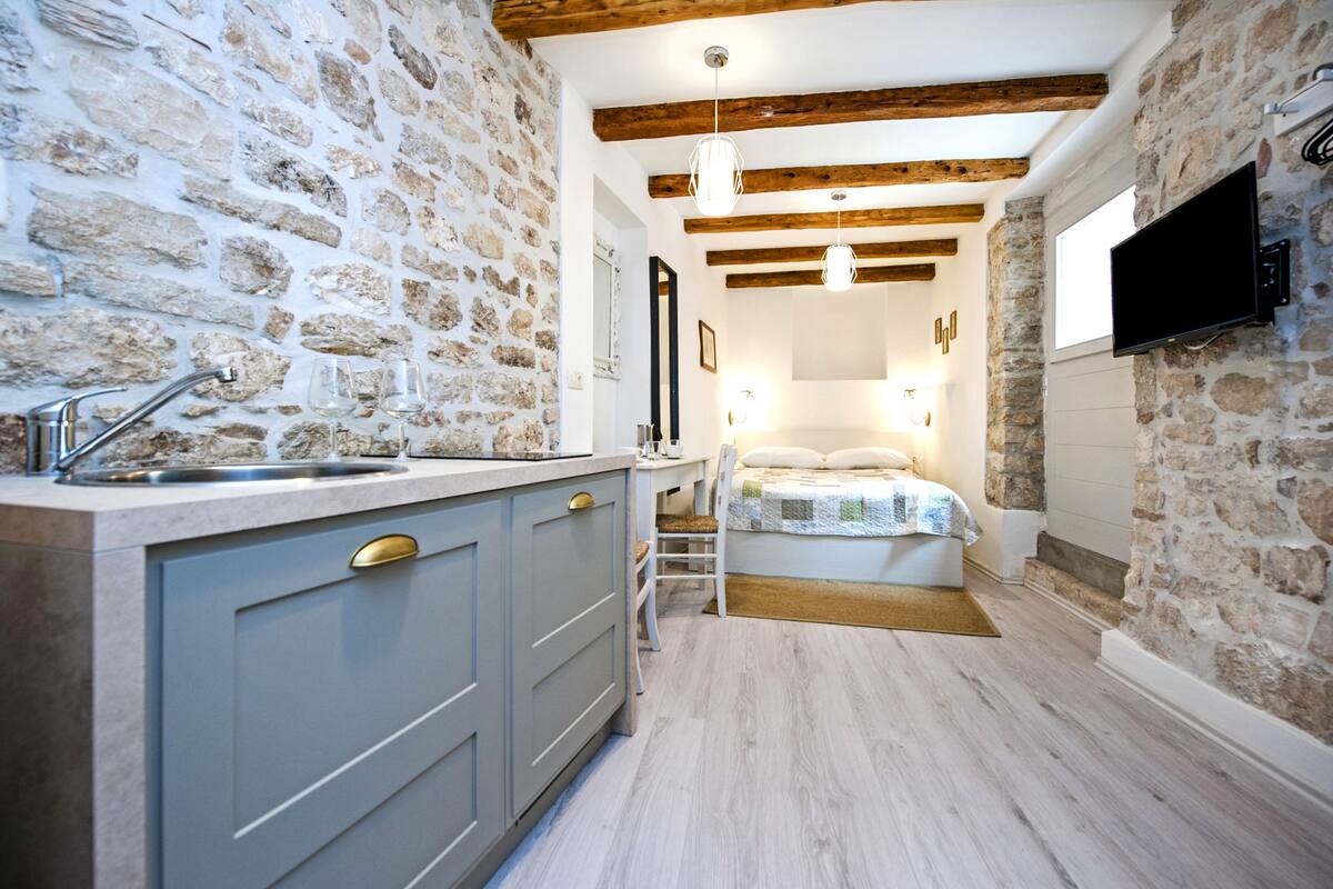 Een compacte kamer met een stenen muur, houten balken, een bed, een televisie en een kitchenette.