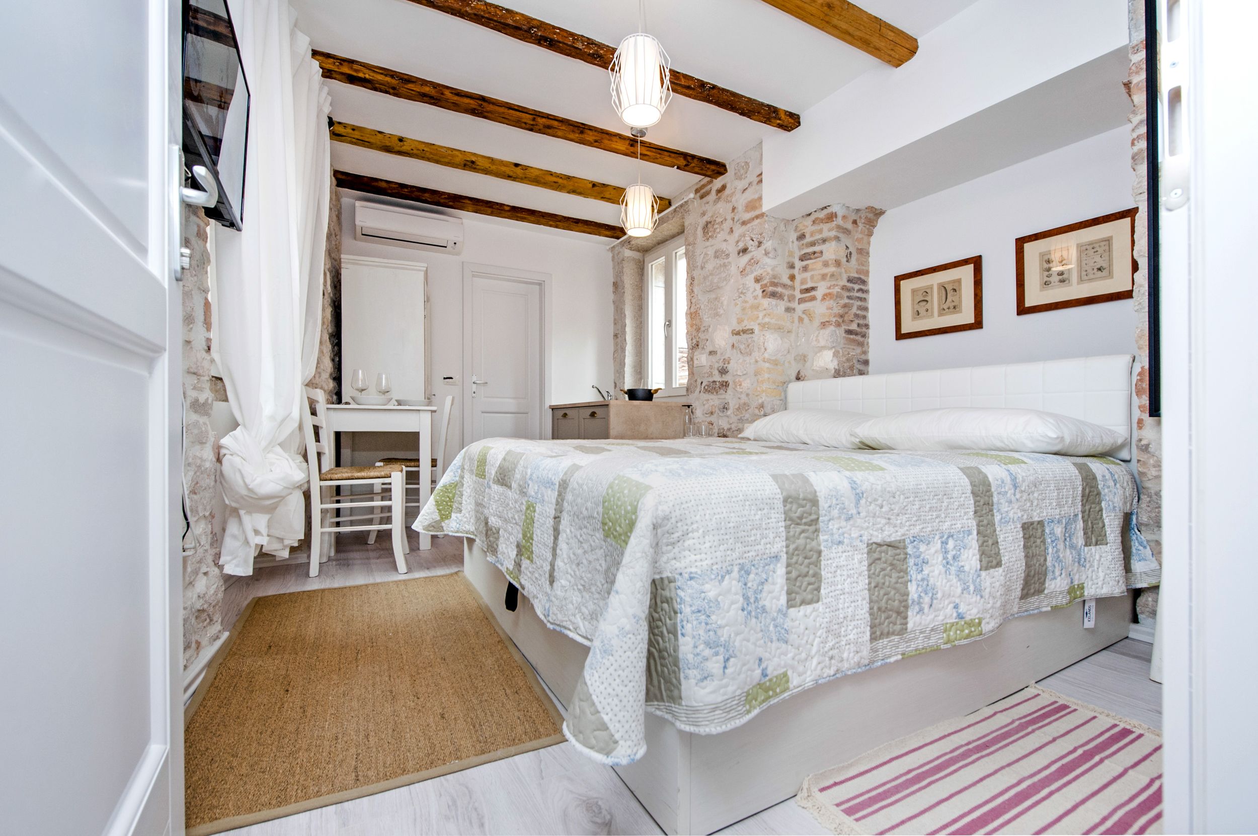 Un dormitorio con una cama, un escritorio y una silla. Hay una ventana, una puerta y una pared de piedra.